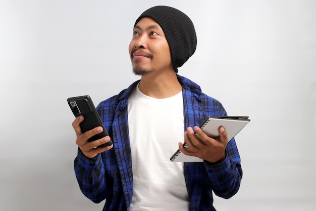 Excited Asian man holds a phone and a notebook looking aside at empty copy space