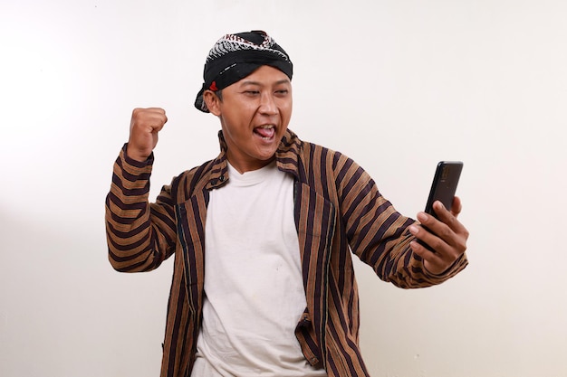 Excited asian man in javanese costume holding a cell phone while clenching his hand