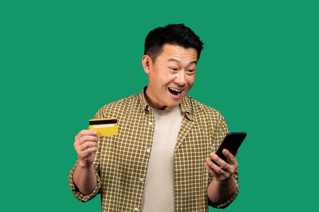 Возбужденный азиатский мужчина использует мобильный телефон и кредитную карту для покупок в Интернете, наслаждаясь приложением для электронной коммерции