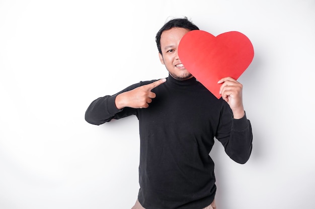 Excited Asian man wearing black shirt pointing at the copy space beside him while holding a big red heartshaped paper isolated by white background