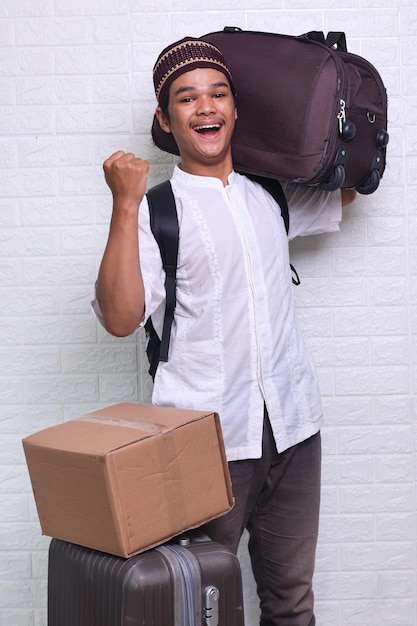 Excited Asian muslim man carrying travel suitcase and cardboard giving yes gesture Going mudik or