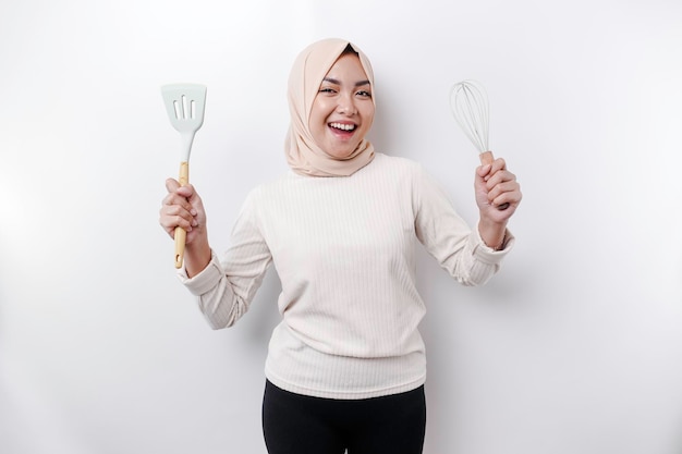 Excited Asian Muslim woman wearing a headscarf holding cooking ware and smiling isolated by white background