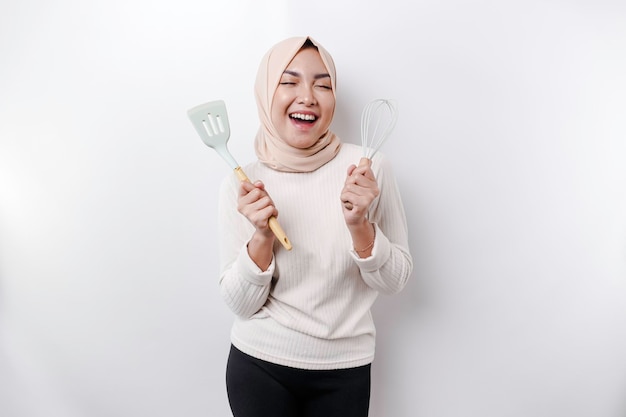 Excited Asian Muslim woman wearing a headscarf holding cooking ware and smiling isolated by white background