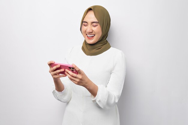 Excited Asian Muslim woman wearing hijab play games with a mobile phone playing a racing app on gadget isolated on white background studio portrait People religious lifestyle concept