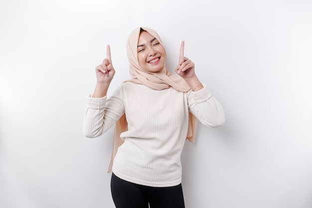 Excited Asian Muslim woman wearing hijab pointing at the copy space above her isolated by white background