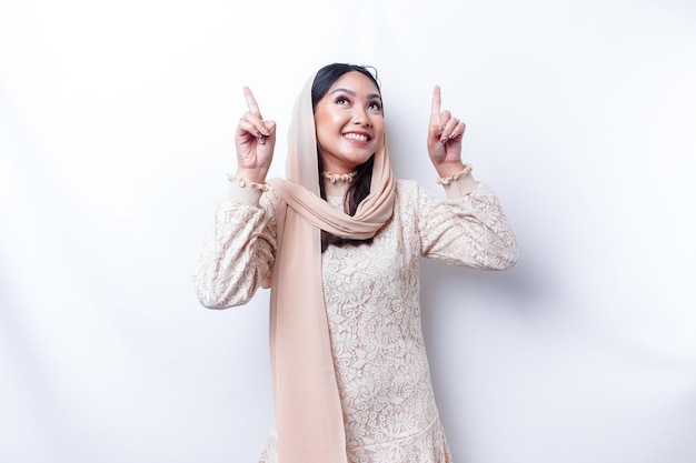 히잡을 쓴 흥분한 아시아 무슬림 여성이 흰색 배경에 의해 고립된 그녀 위의 복사 공간을 가리키고 있습니다.