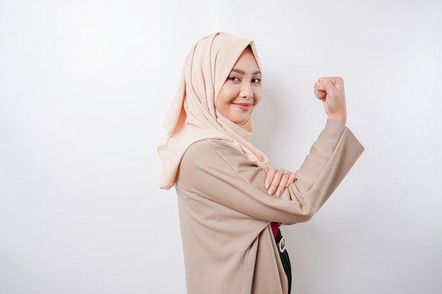 Excited Asian Muslim woman wearing a hijab showing strong gesture by lifting her arms and muscles smiling proudly