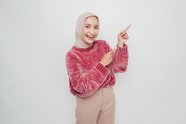 분홍색 스웨터와 히잡을 쓴 흥분한 아시아 무슬림 여성이 흰색 배경에 의해 고립된 그녀 옆에 있는 복사 공간을 가리키고 있습니다.