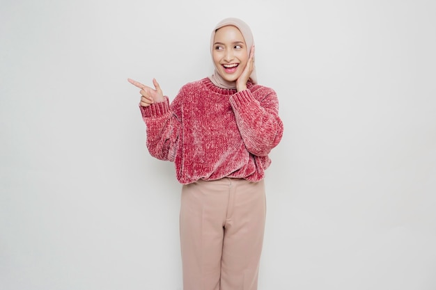 Excited Asian Muslim woman wearing pink sweater and hijab pointing at the copy space beside her isolated by white background