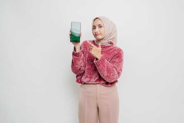 Excited Asian Muslim woman wearing pink sweater and hijab pointing at the copy space beside her while holding her phone isolated by white background