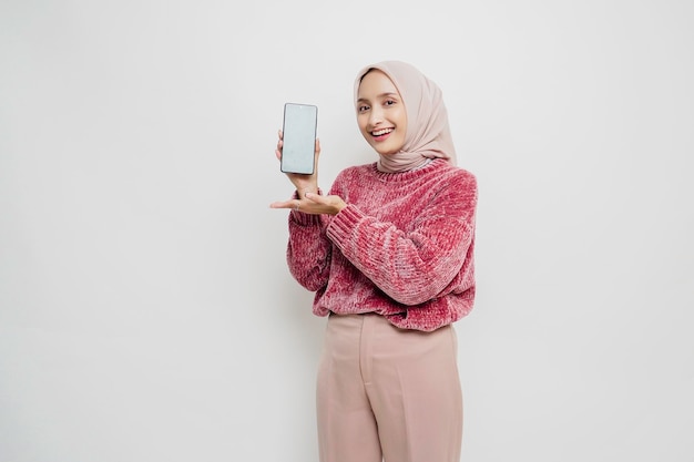 Excited Asian Muslim woman wearing pink sweater and hijab pointing at the copy space beside her while holding her phone isolated by white background