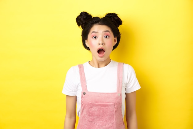 Возбужденная азиатская девушка-подросток отвисает челюстью, задыхается от удивления открытым ртом, впечатленно смотрит в камеру, проверяет летнее промо-предложение, стоит на желтом.