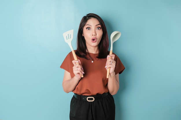 Excited Asian woman holding cooking ware and smiling isolated by blue background