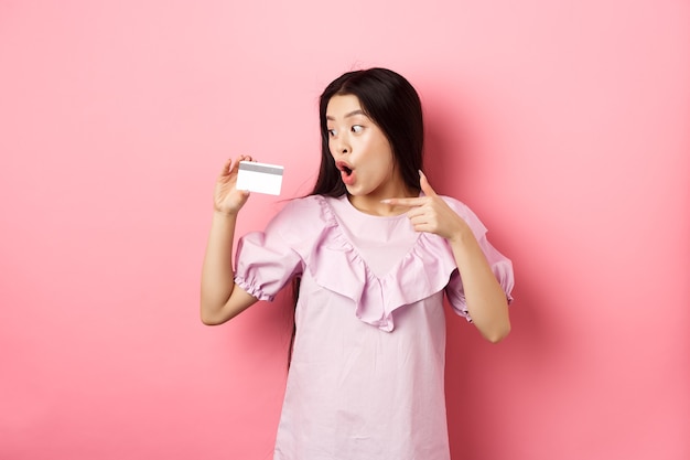 Excited asian woman pointing and showing plastic credit card, gasping amazed, concept of shopping.