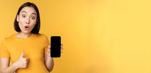 Excited asian woman showing mobile phone screen thumbs up like smth good recommending smartphone application standing over yellow background