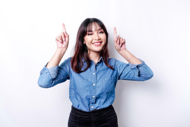 Excited Asian woman wearing blue shirt pointing at the copy space on top of her isolated by white background