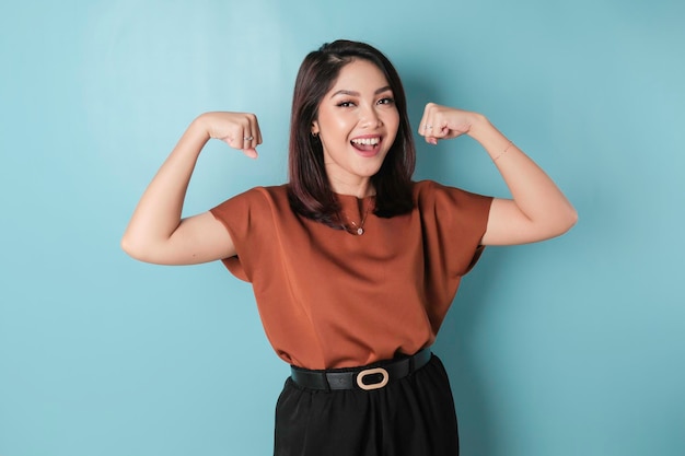 갈색 셔츠를 입은 흥분한 아시아 여성이 자랑스럽게 미소 짓는 팔과 근육을 들어 올려 강한 제스처를 보여줍니다.