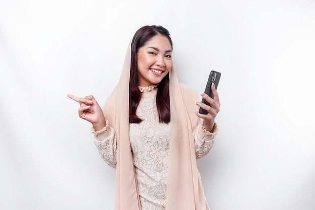 Excited Asian woman wearing hijab pointing at the copy space beside her while holding her phone isolated by white background