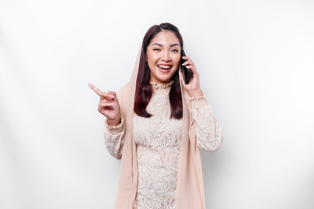 Excited Asian woman wearing hijab pointing at the copy space beside her while holding her phone isolated by white background