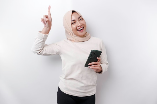 Excited Asian woman wearing hijab pointing at the copy space on top of her while holding her phone isolated by white background