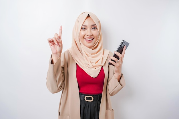 Excited Asian woman wearing hijab pointing at the copy space on top of her while holding her phone isolated by white background