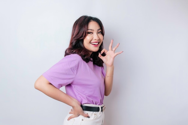 Excited Asian woman wearing a lilac purple tshirt giving an OK hand gesture isolated by a white background