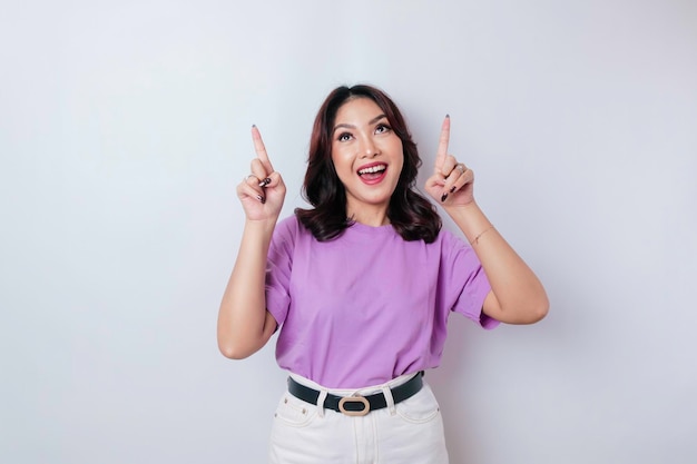 Excited Asian woman wearing lilac purple tshirt pointing at the copy space upside her isolated by white background