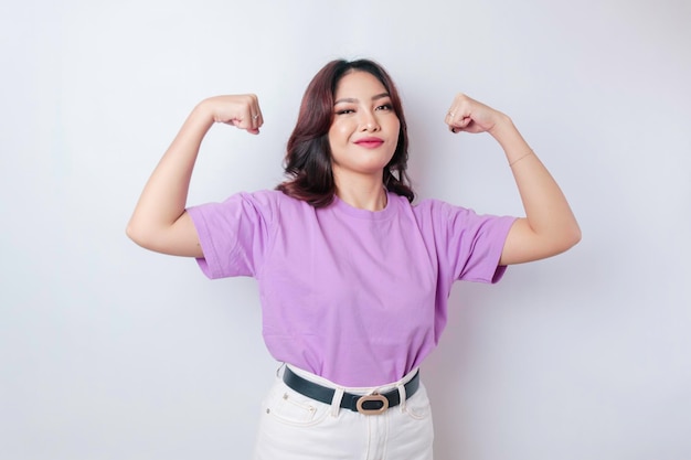 腕と筋肉を誇らしげに持ち上げて強いジェスチャーを示すライラックパープルのTシャツを着た興奮したアジア人女性