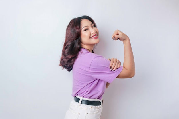 Excited Asian woman wearing a lilac purple tshirt showing strong gesture by lifting her arms and muscles smiling proudly