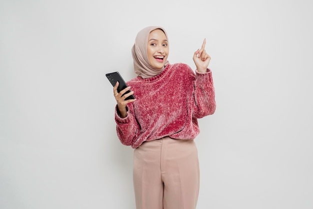 Excited Asian woman wearing pink sweater and hijab pointing at the copy space on top of her while holding her phone isolated by white background