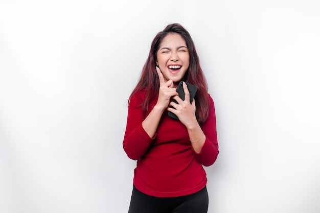 Excited Asian woman wearing red top pointing at the copy space on top of her while holding her phone isolated by white background