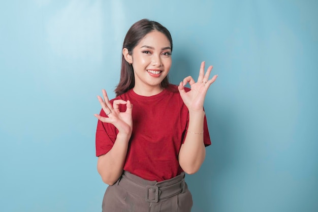 Excited Asian woman wearing a red tshirt giving an OK hand gesture isolated by a blue background