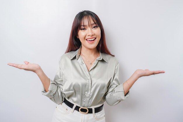 Excited Asian woman wearing sage green shirt pointing at the copy space beside her isolated by white background