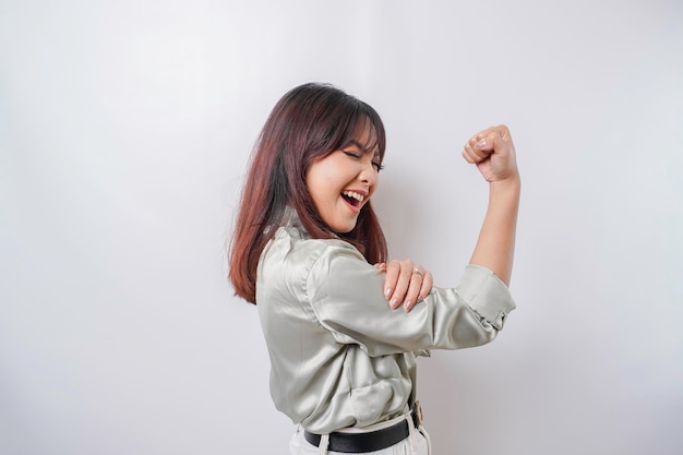 Excited Asian woman wearing a sage green shirt showing strong gesture by lifting her arms and muscles smiling proudly