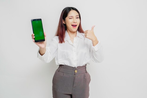 Excited Asian woman wearing white shirt gives thumbs up hand gesture of approval while showing copy space on her phone