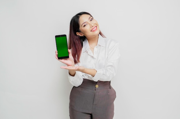 Excited Asian woman wearing white shirt pointing at the copy space beside her while holding her phone isolated by white background