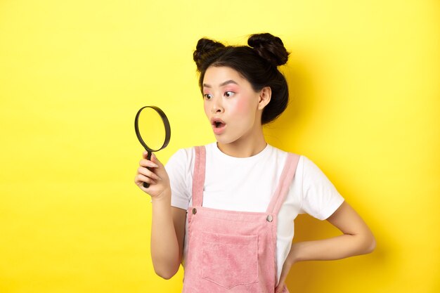 Photo excited asian woman with bright glamour makeup, look through magnifying glass amazed, staring aside, standing on yellow