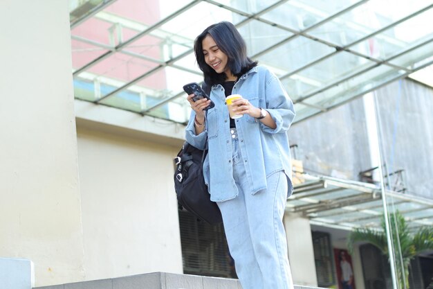 アジアの興奮した若い女性は ⁇ スマートフォンのアプリを使用し ⁇ モールの屋外の階段でコーヒーを飲みながら笑っています ⁇ 