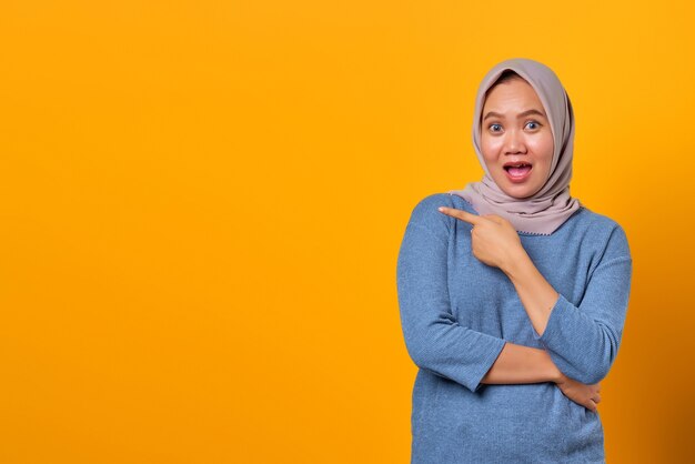 Excited attractive asian woman pointing finger at empty space over yellow background