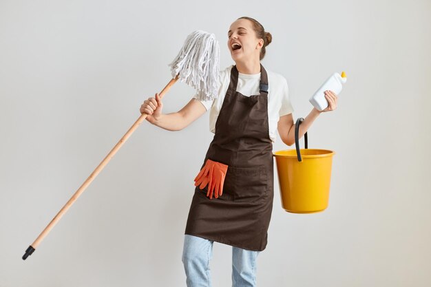 Возбужденная привлекательная домохозяйка в белой футболке и коричневом фартуке держит оборудование для уборки дома, держит желтое ведро и швабру и поет как микрофон