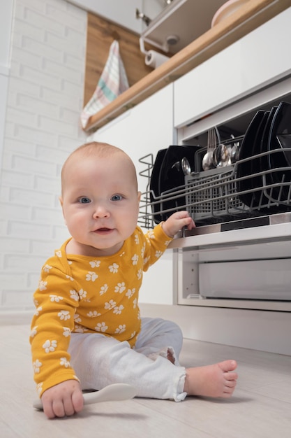 Excited baby girl plays with spoon at dishwasher in kitchen