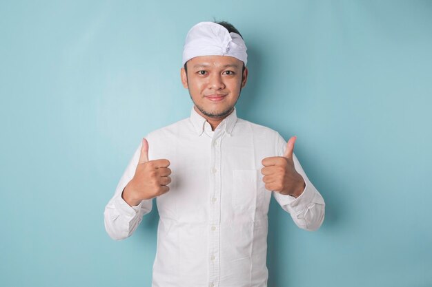 Excited Balinese man wearing udeng or traditional headband and white shirt gives thumbs up hand gesture of approval isolated by blue background