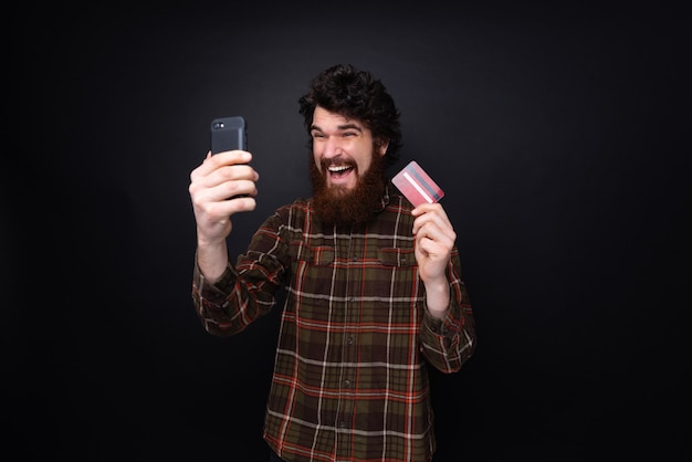 Excited bearded guy holding mobile phone and credit card, standing over dark wall background