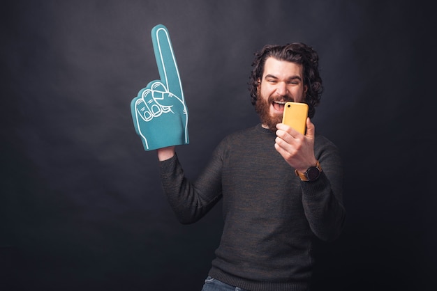 Excited bearded handsome man pointing with fan glove and looking at yellow smartphone