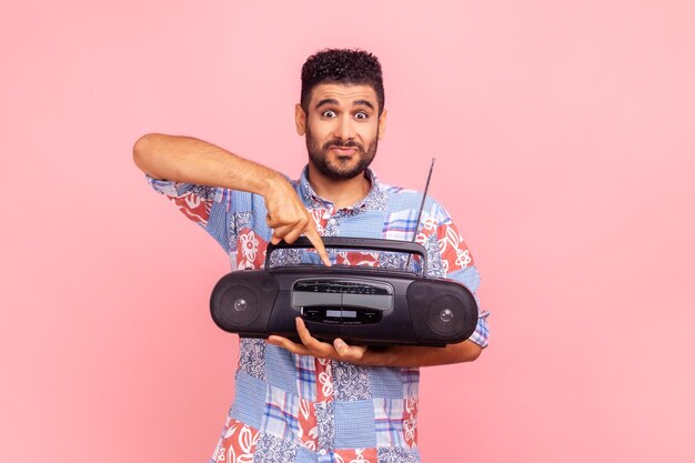 Excited bearded man in blue casual style shirt holding in hand black tape recorder pushing play button looking at camera with funny expression indoor studio shirt isolated on pink background