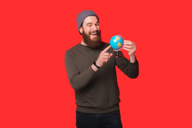 Excited bearded man is holding a globe and pointing on it.