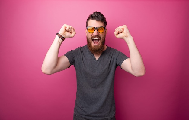Excited bearded man, screaming and celebrating with rised hands, standing over pink background