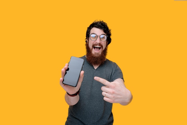 Excited bearded man wearing glasses, is pointing at smartphone on yellow