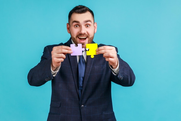 Excited bearded man wearing official style suit holding and showing two jigsaw pieces matching and connecting puzzles solving problems creativity Indoor studio shot isolated on blue background