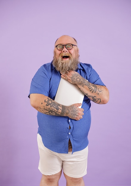 Photo excited bearded obese man in tight blue shirt with glasses embraces modern laptop standing on purple background in studio